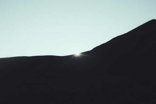 Capture of sunlight peeking over a hill silhouette under a clear sky at dawn, serene and tranquil.