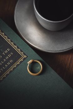 A close-up of an elegant golden ring placed on a book beside a cup of coffee on a saucer.