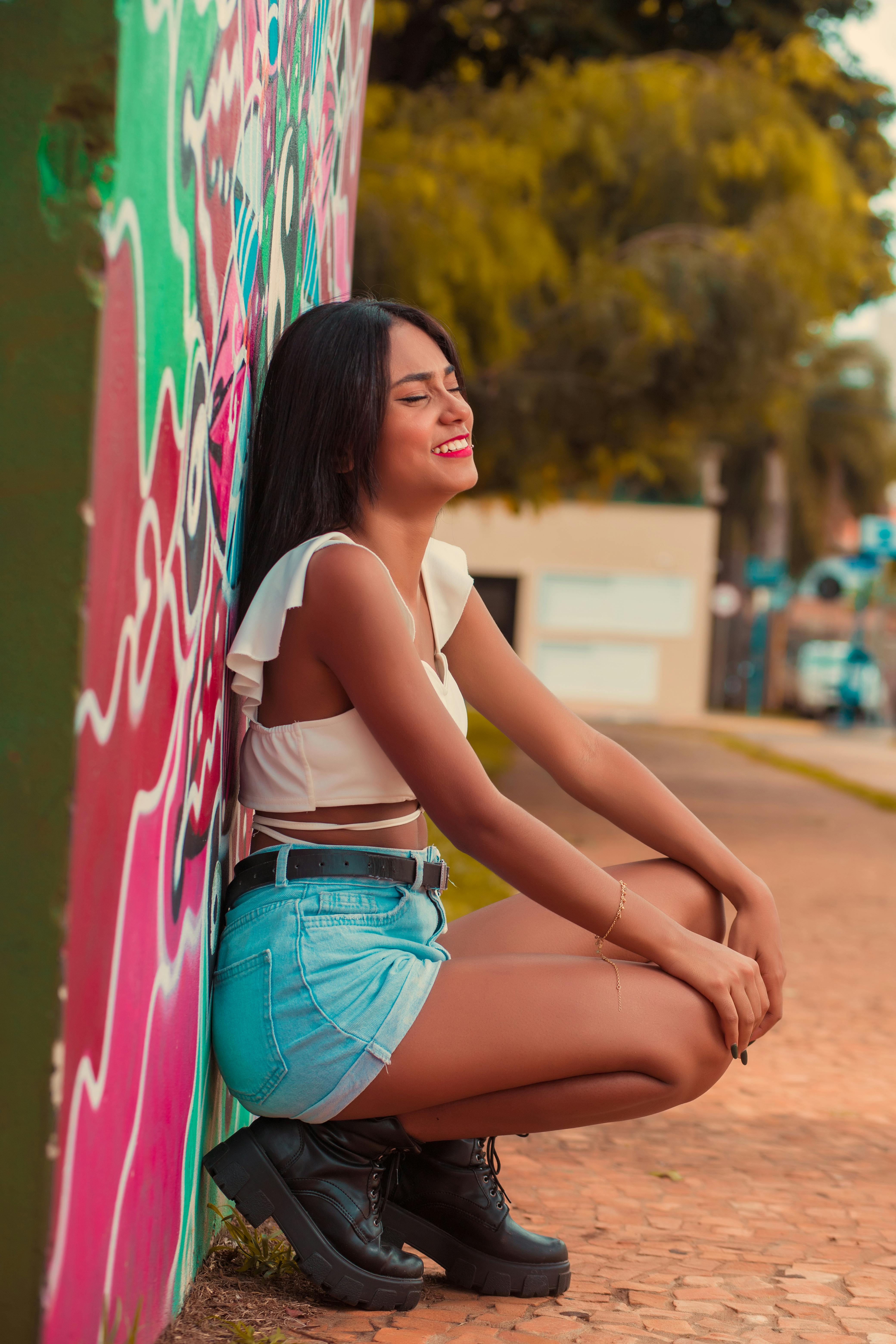 A stylish woman in casual attire smiles while leaning against a graffiti-covered wall outdoors.