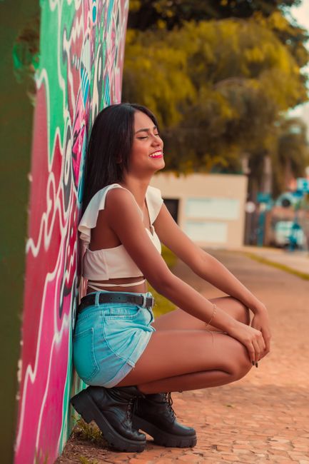 A stylish woman in casual attire smiles while leaning against a graffiti-covered wall outdoors.