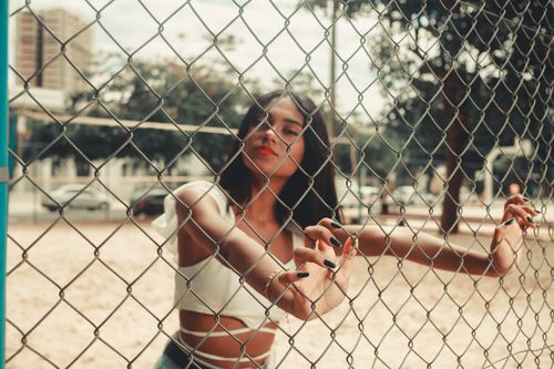 Free Stylish young woman in summer attire poses behind wire mesh fence outdoors. Stock Photo