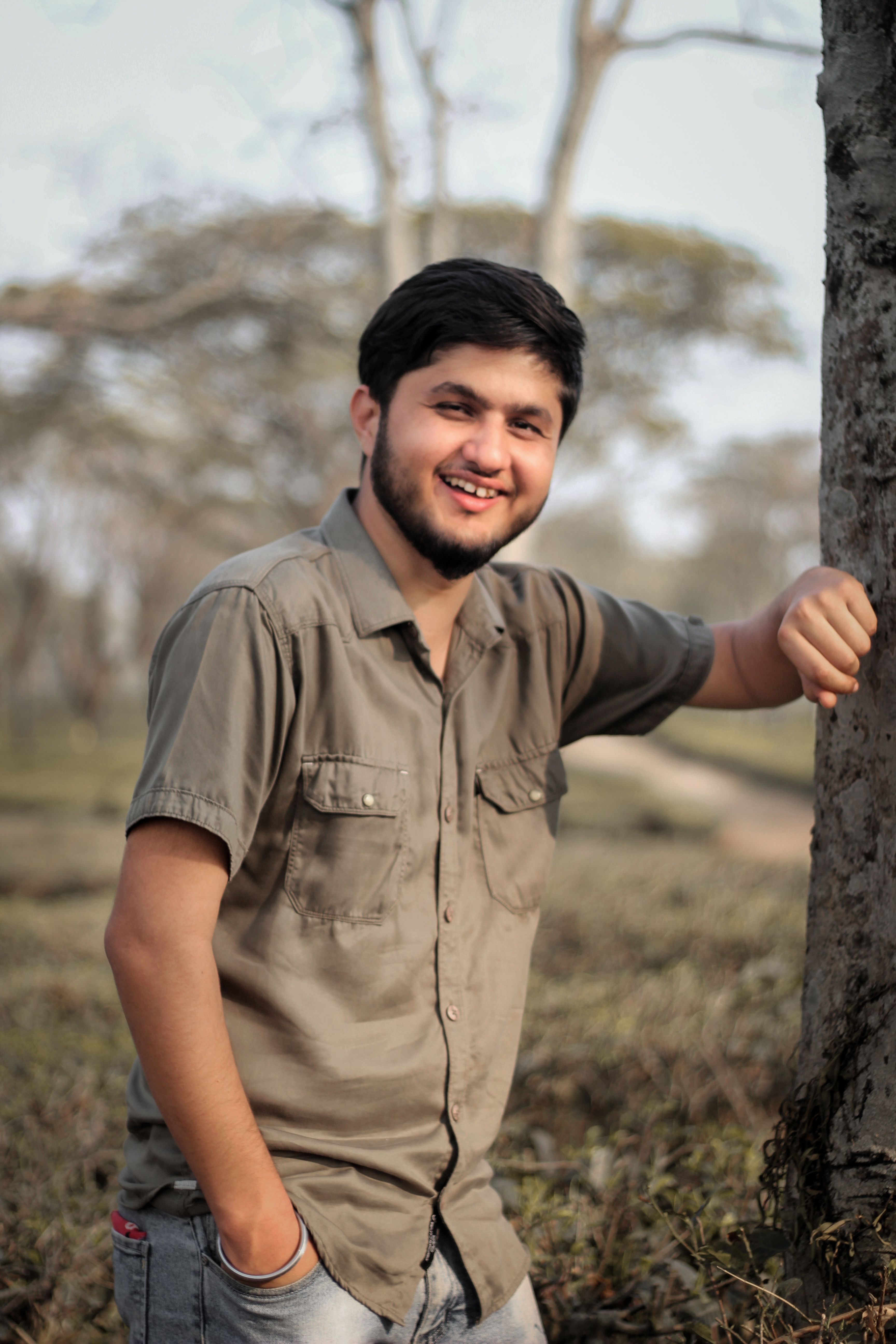 A smiling young man leans against a tree in a casual outfit, enjoying a sunny day in the park.