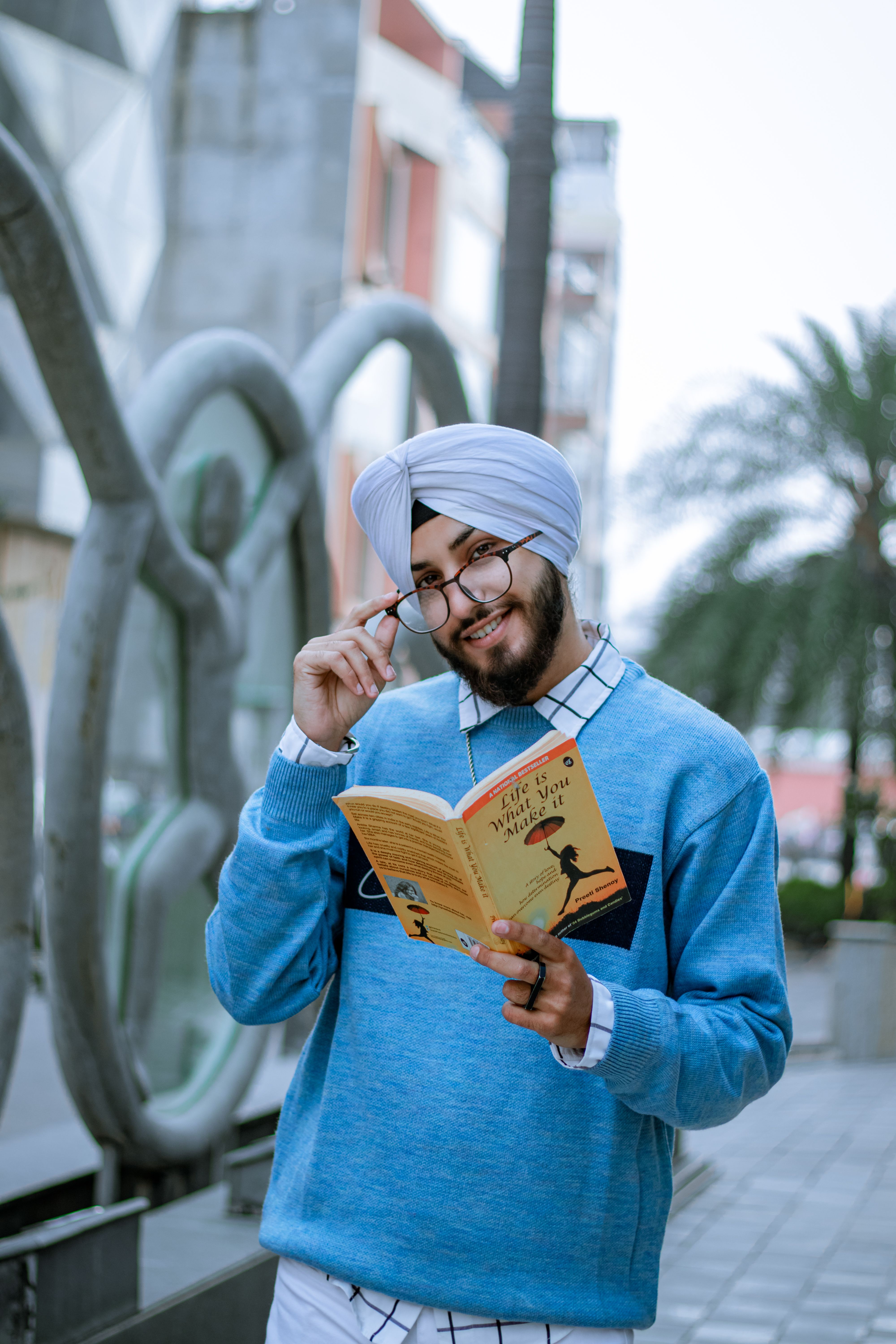 Man with turban and glasses reading a book on a city street, smiling.