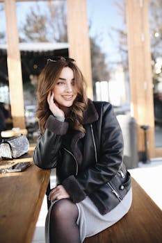 Young woman in a leather jacket smiling at an outdoor café table with accessory details.