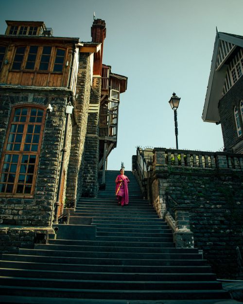 Free A woman in a saree walks down stairs near a historic stone building on a sunny day. Stock Photo