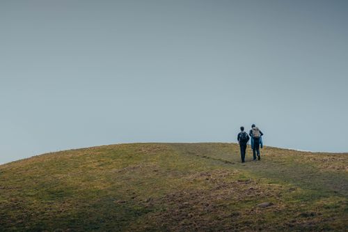 Free Two hikers with backpacks walking up a grassy hill on a clear day outdoors. Stock Photo