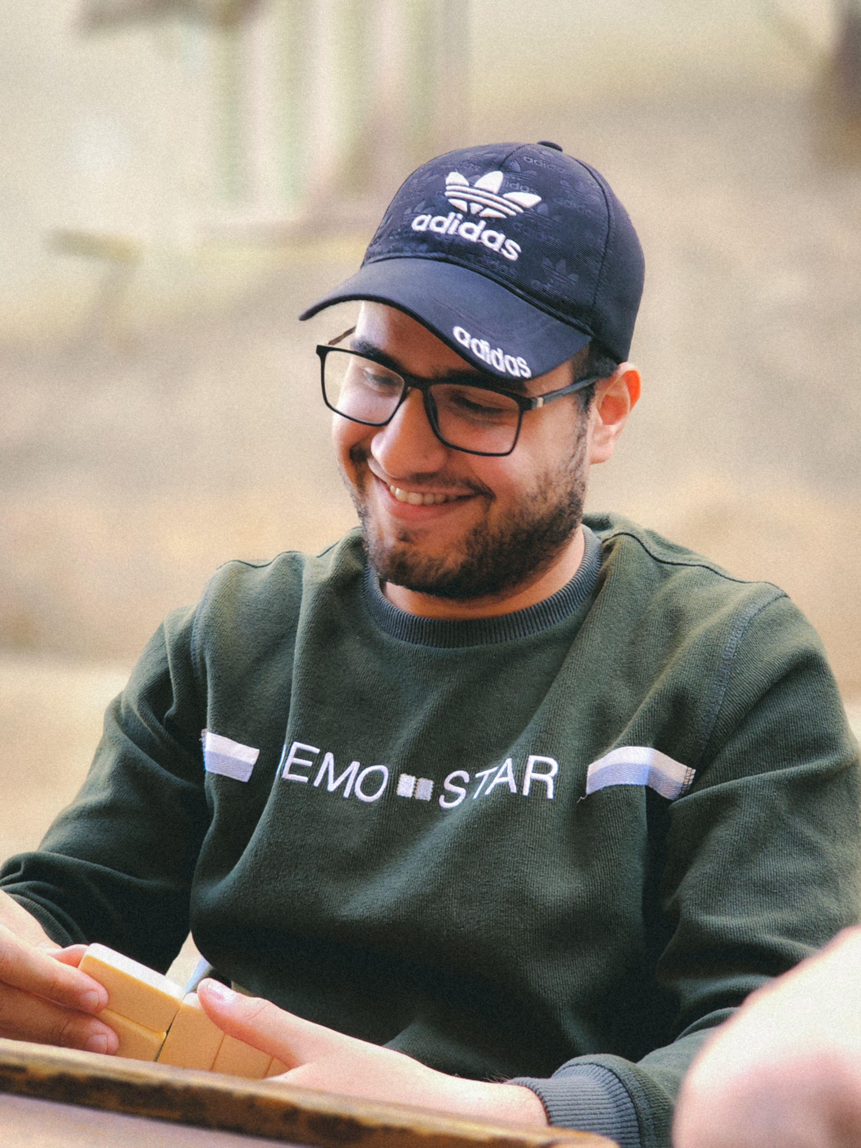 A cheerful man wearing a cap and glasses playing dominoes outside, dressed casually.