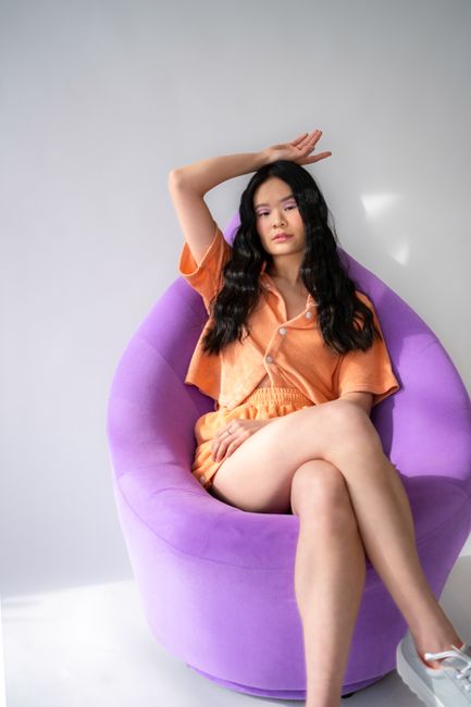 Elegant Asian woman sits on a purple chair, showcasing vibrant fashion in a studio setting.