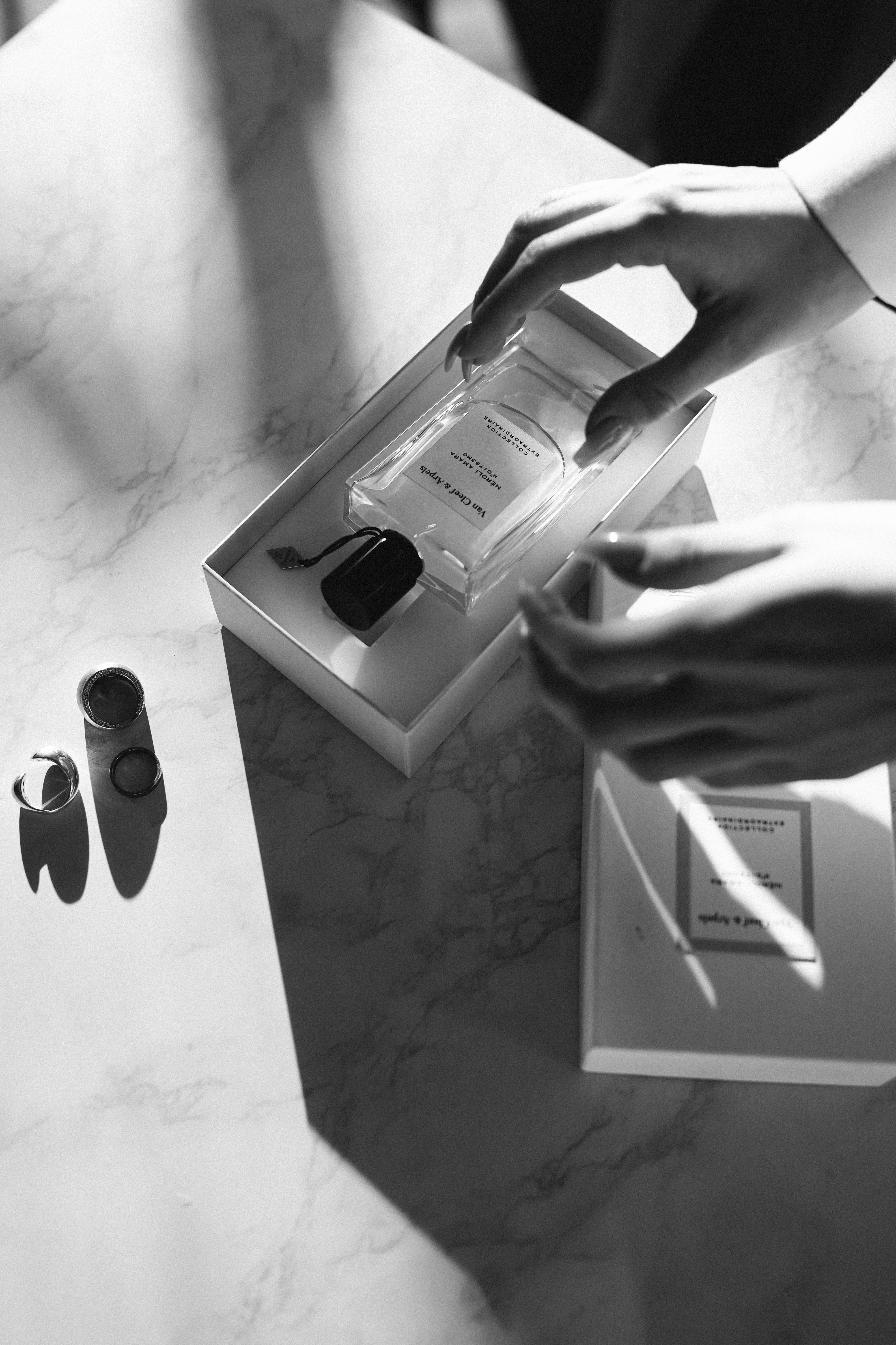 Monochrome image of hands opening a perfume bottle box on a marble surface.