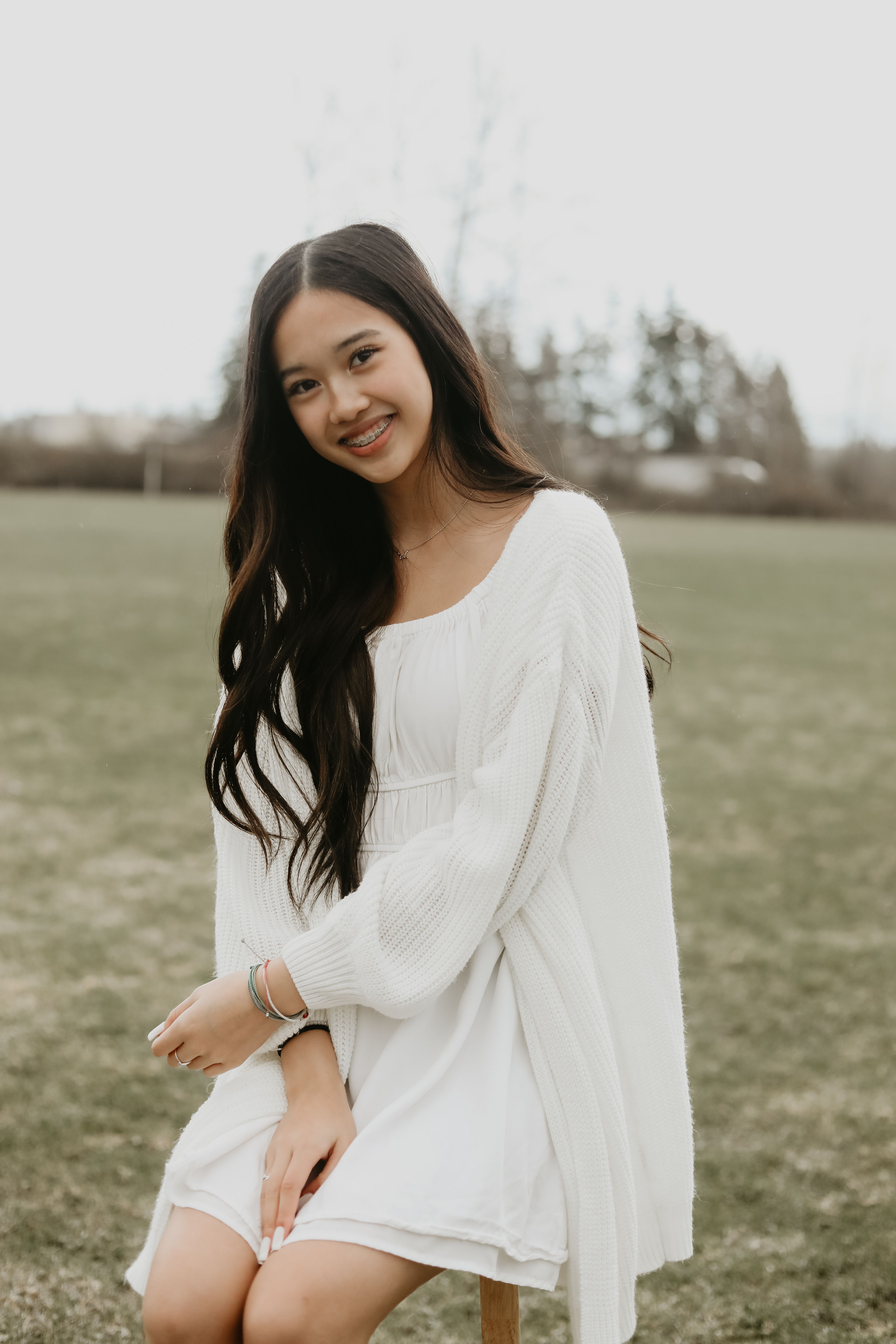 Beautiful Asian woman in a white dress smiling outdoors, sitting in an open field.