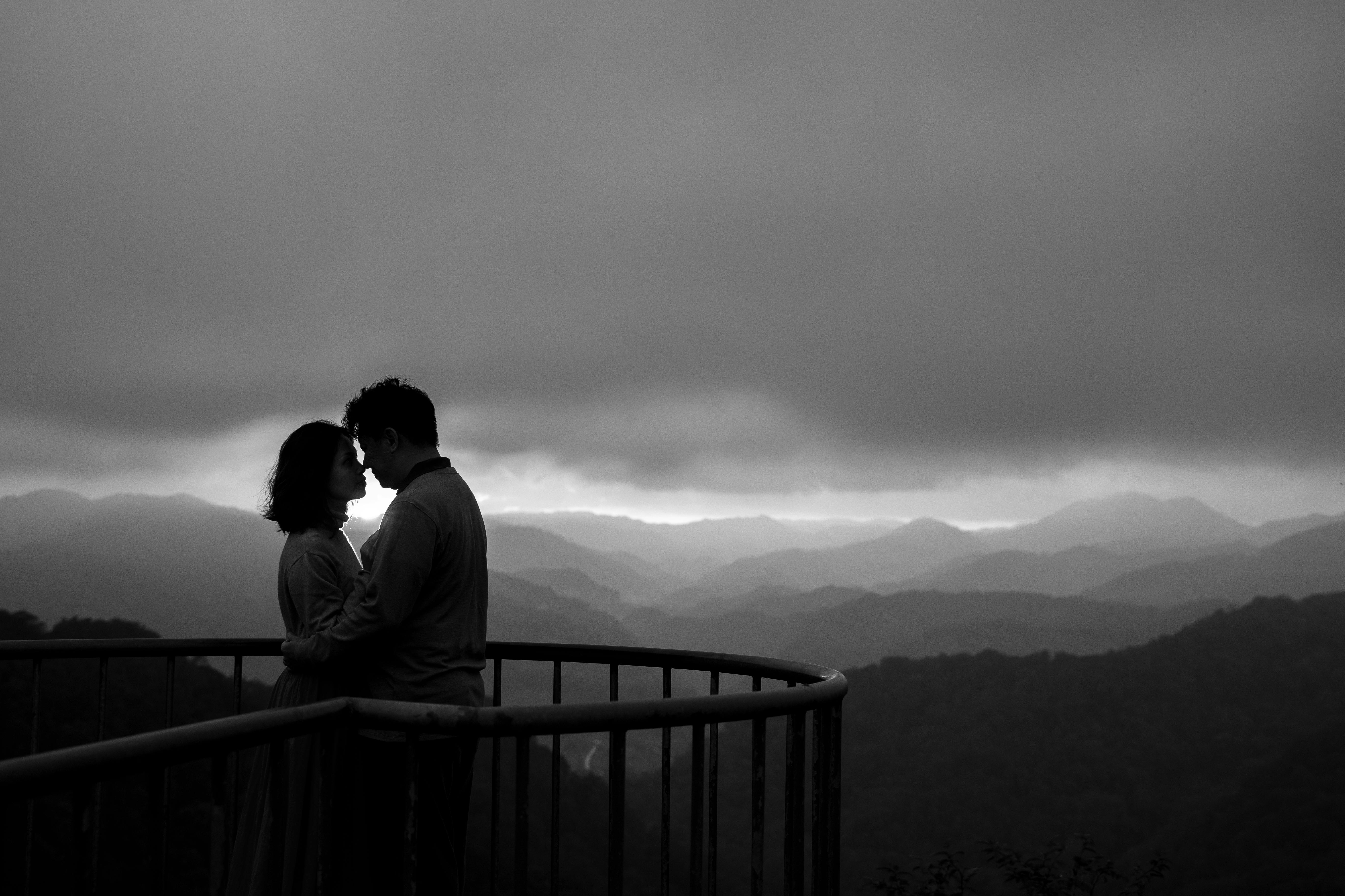 Silhouette of a couple embracing on a scenic viewpoint with clouds over mountains.