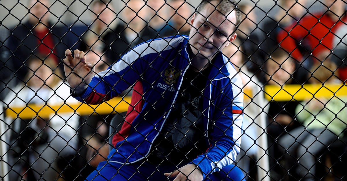 Athlete in tracksuit focuses through a fence at a match with spectators in background.