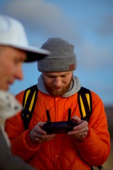 A man with a beanie and orange jacket focuses on controlling a drone outdoors.