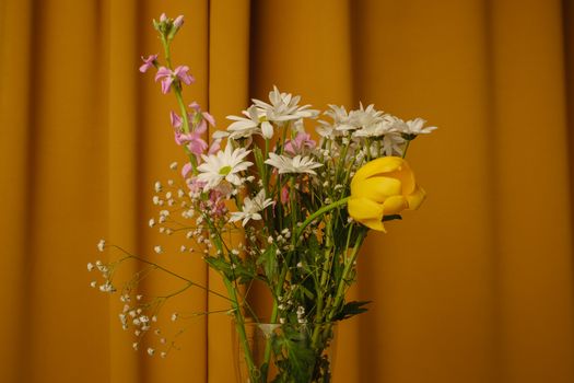 A vivid still life of a floral bouquet in a glass vase set against a warm yellow curtain backdrop.