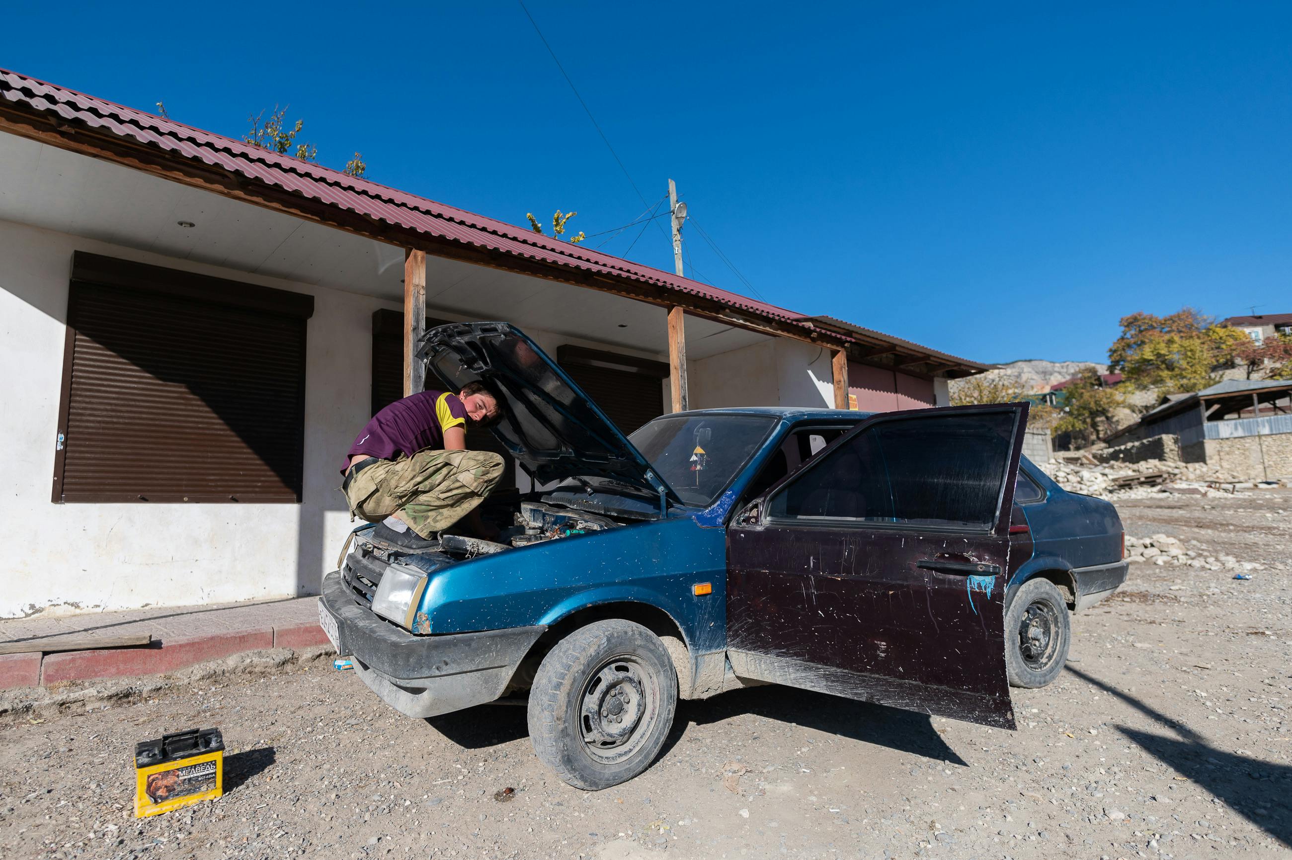 Adult male repairing a car outdoors in a rural setting, near a house.