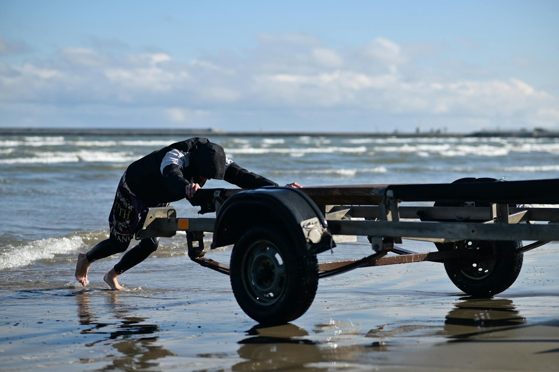 Barefoot man struggles to push trailer on a wave-kissed beach under a bright sky.