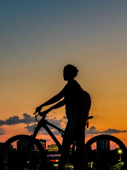 Free Silhouette of a woman with a bicycle against a vibrant sunset sky, urban backdrop. Stock Photo