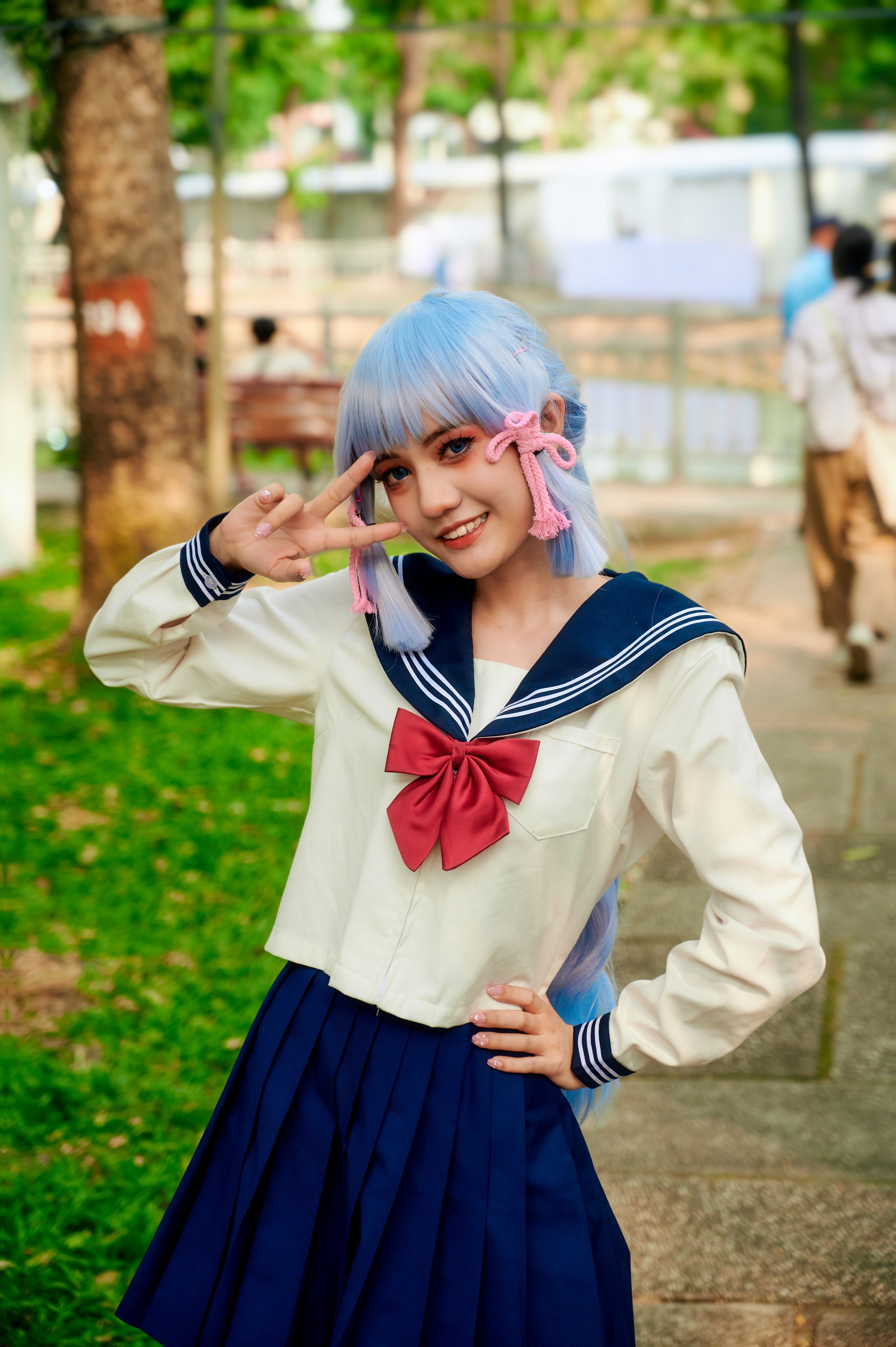 Teenage girl with blue hair in Japanese school uniform posing playfully outdoors in a park.