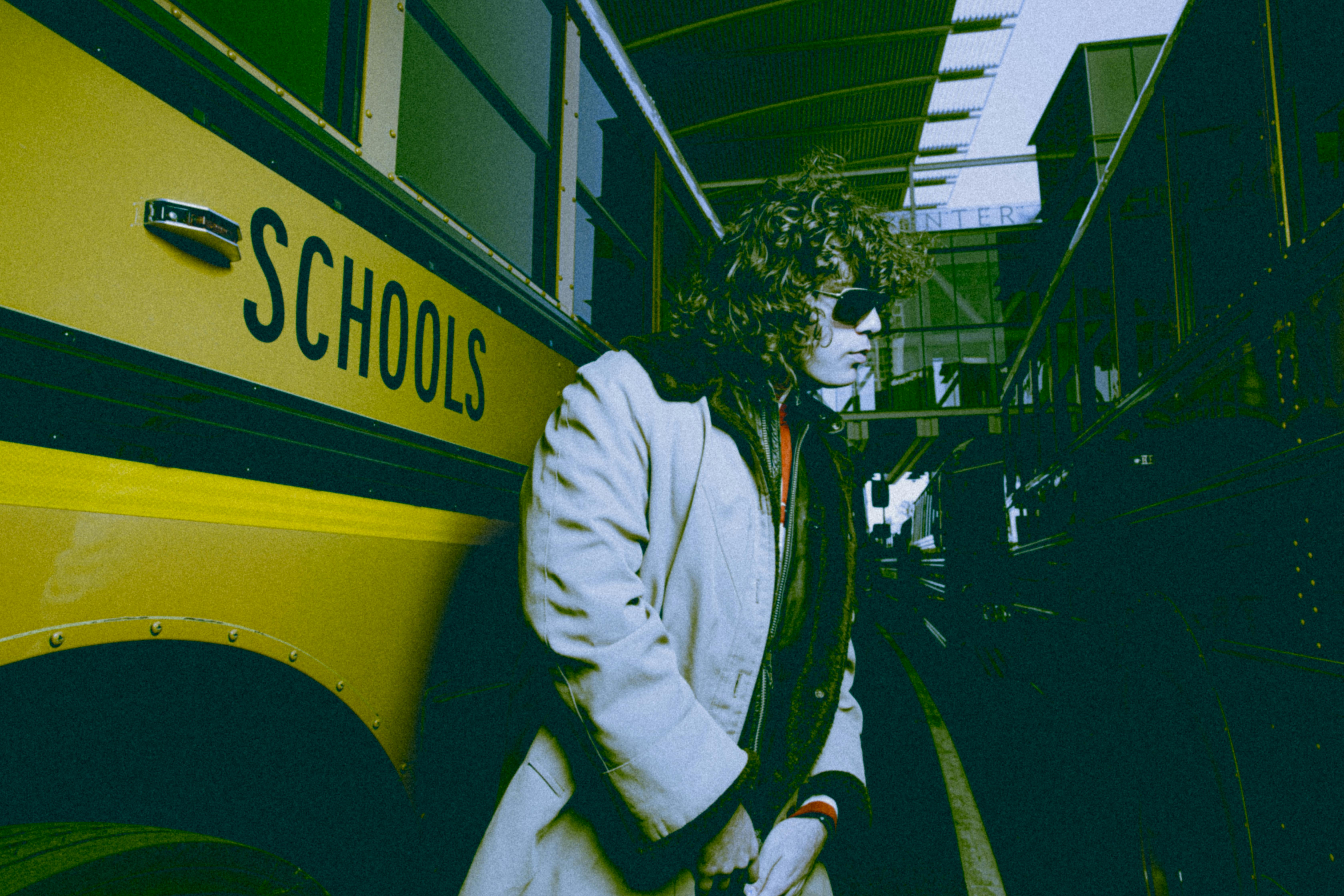 Man in coat and sunglasses standing next to a school bus in an urban setting.