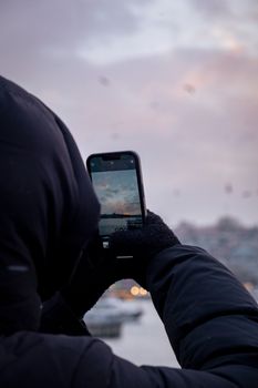 Person in winter attire photographs a sunrise with a smartphone outdoors.