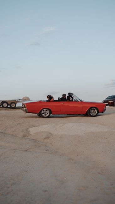 Vintage red convertible with passengers in a serene desert setting at dawn, evoking a retro travel vibe.