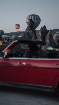 Colorful hot air balloons above unique rock formations at twilight with a vintage convertible in the foreground.