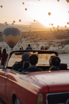 A scenic view of hot air balloons at sunrise in Cappadocia with a vintage car in the foreground.