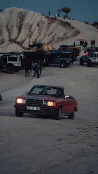 Classic red Mercedes cabriolet navigating a scenic desert landscape, surrounded by off-road vehicles and travelers.