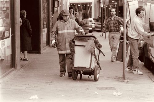 Free A garbageman pushes a cart down a busy urban street, capturing everyday life in sepia tones. Stock Photo
