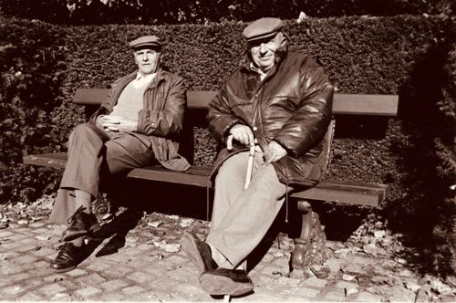Free Sepia-toned image of two elderly men sitting on a sunlit park bench, wearing hats and jackets. Stock Photo