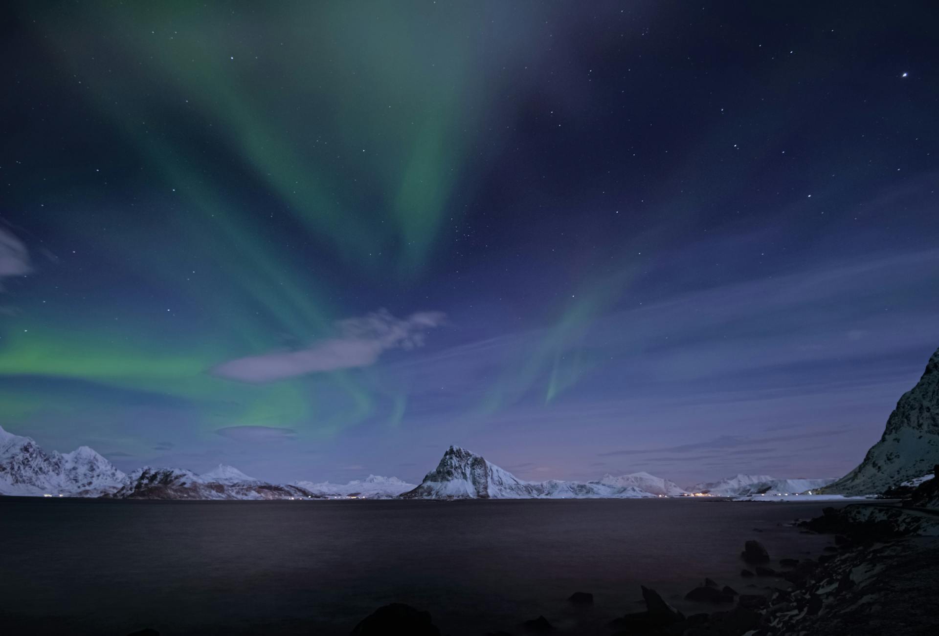 Stunning display of the Northern Lights above snowy mountains and ocean in Lofoten, Norway.