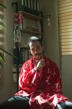 A young man sitting indoors, wrapped in a red festive blanket, enjoying a cozy moment.