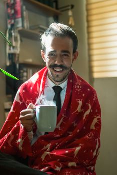 Man wrapped in a festive blanket sipping tea, exuding warmth and comfort indoors.