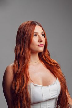 Fashion portrait of a redhead woman with long hair against a gray background.