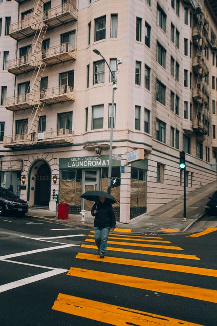 A man crosses a city street under an umbrella by a laundromat on a rainy day.