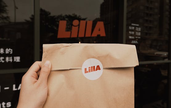 Close-up of a hand holding a brown paper bag with Lilla logo in front of the store's glass facade.