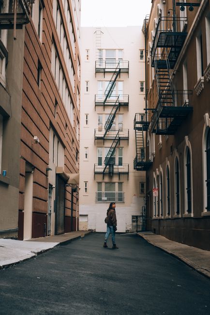 A person walks through a narrow alley lined with buildings and fire escapes at dusk.
