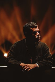 A stylish man with sunglasses on a dimly lit stage in Goiânia, Brazil.