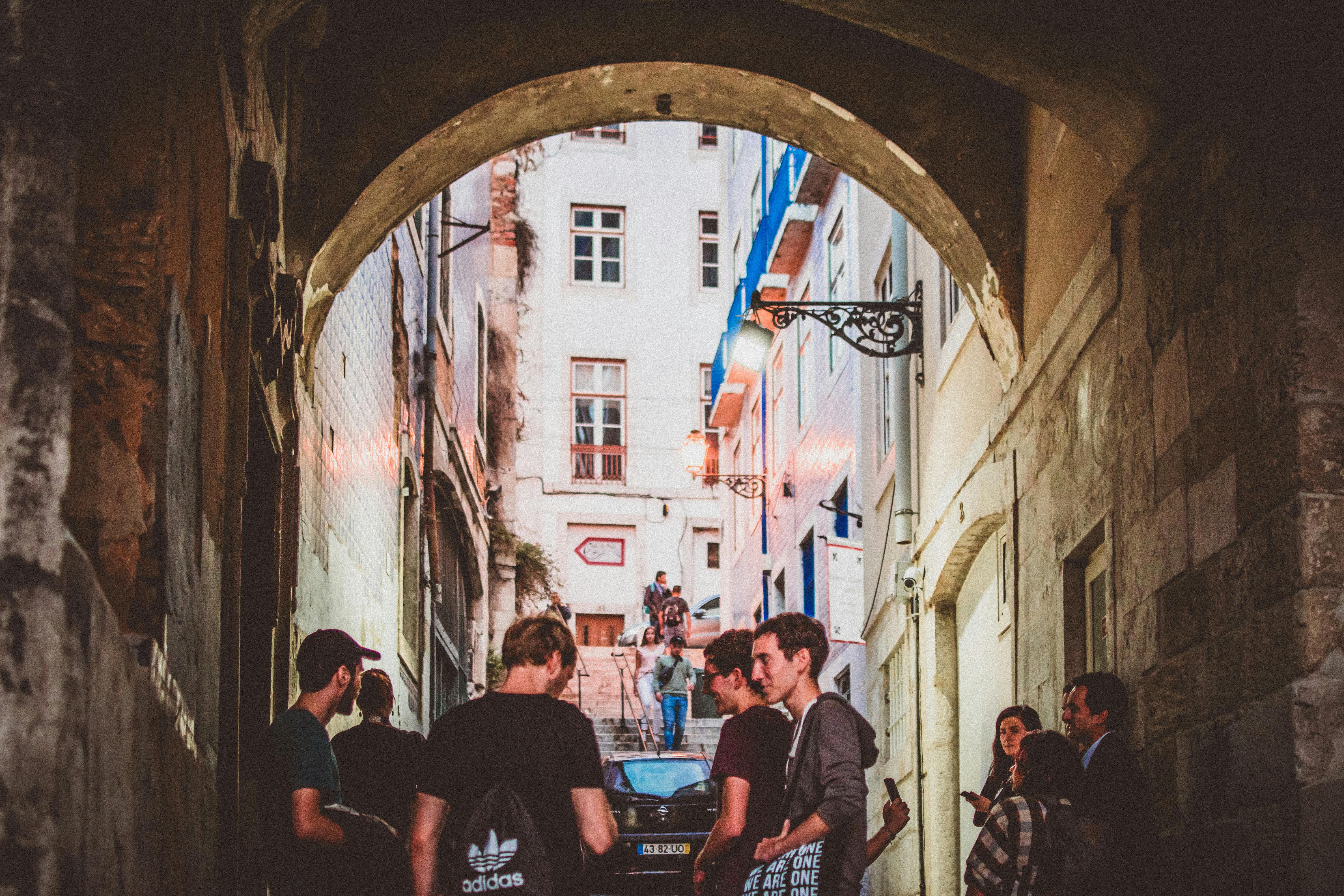 A group of people socializing in a historic Lisbon alleyway, under a stone archway, during daytime.