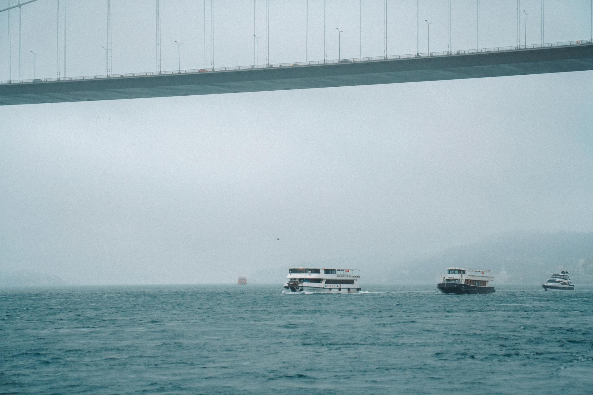 Ferries moving under a large bridge on a foggy day at sea, creating a misty atmosphere.