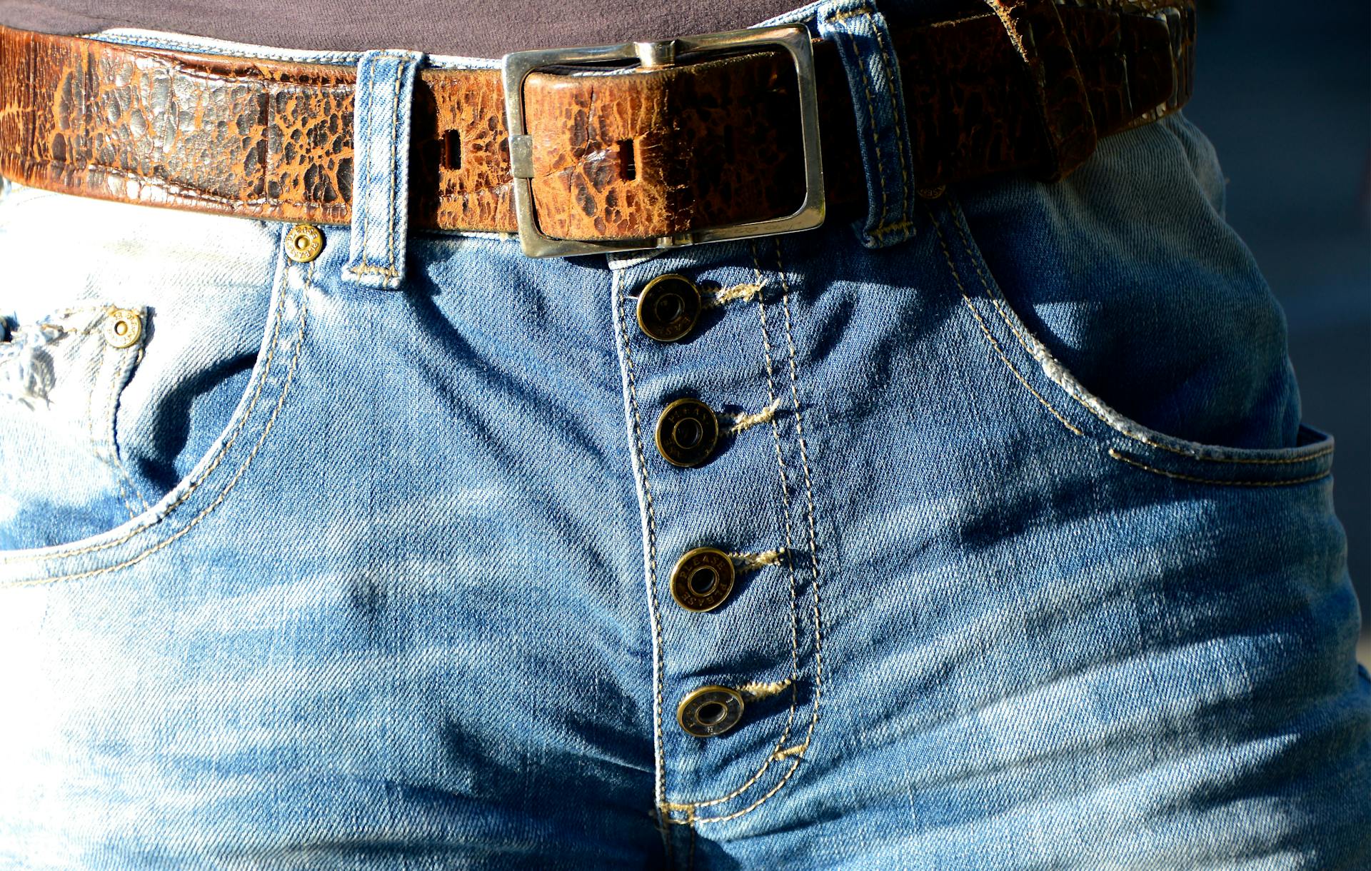 Detailed view of blue denim jeans featuring button fly and brown leather belt.