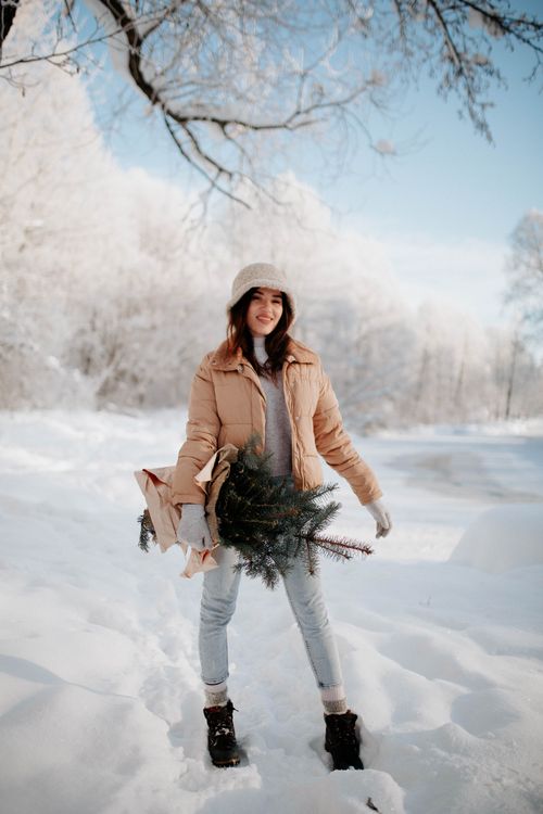 Free Woman in winter outfit carrying evergreen branches in a snowy forest setting. Stock Photo