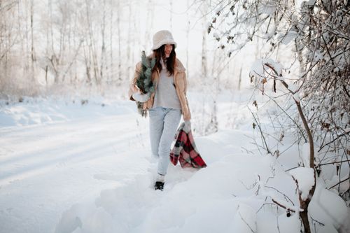Free A young woman in warm clothing walks through a snowy forest carrying a small Christmas tree. Stock Photo
