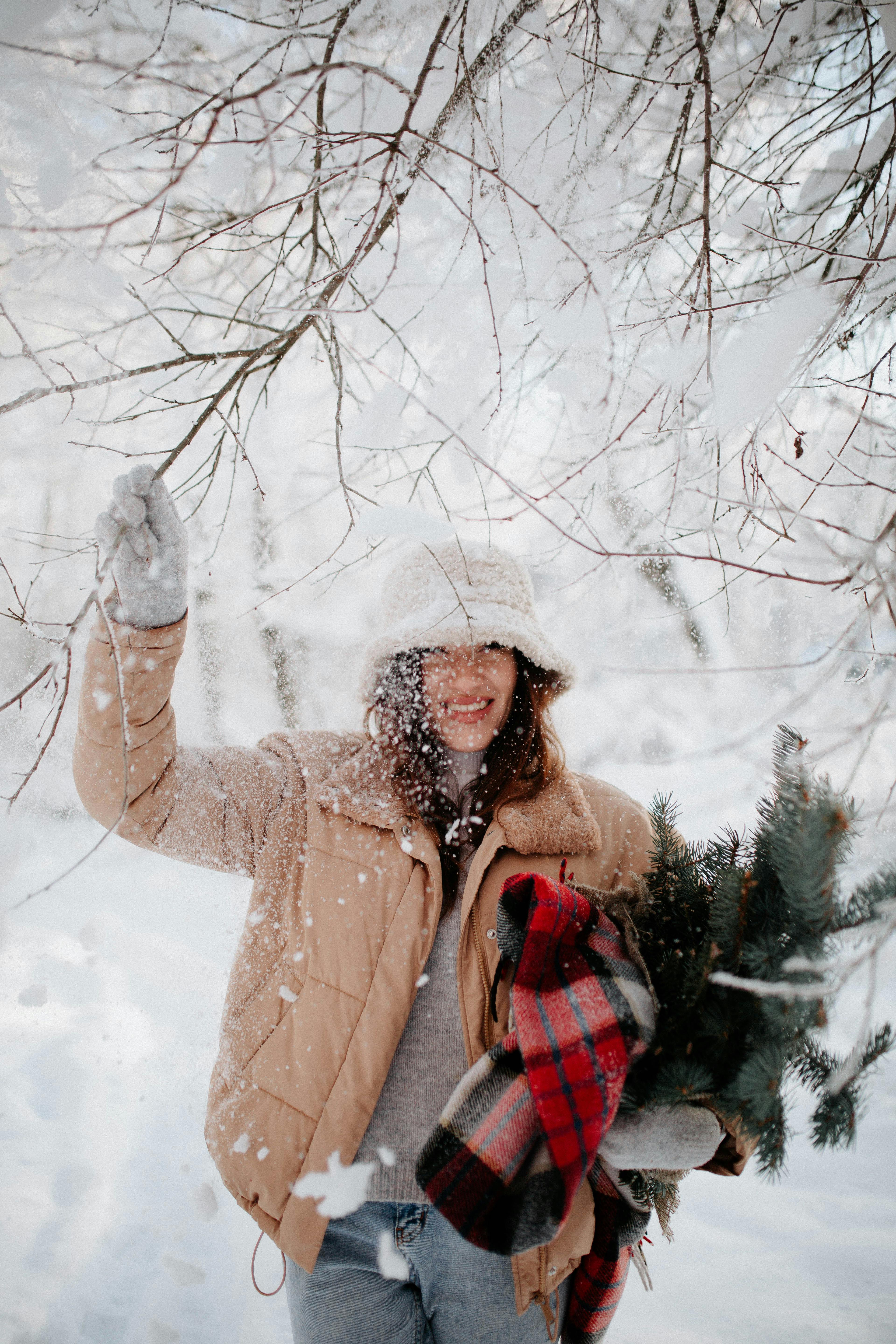 Woman in warm clothing holding evergreen branches, enjoying snowfall outdoors.
