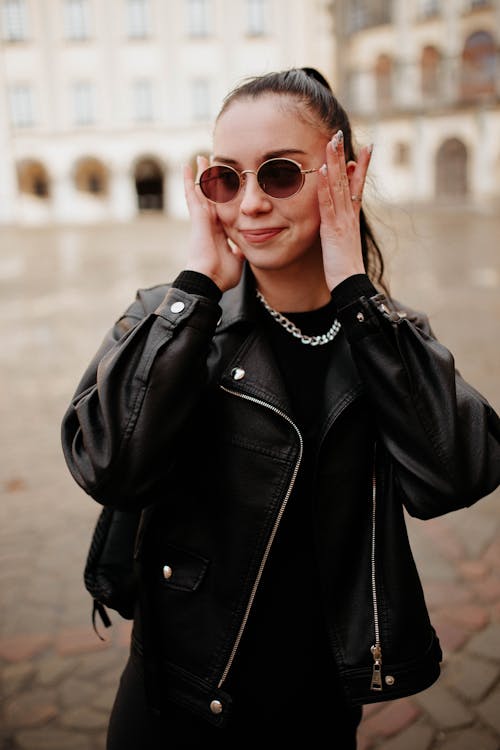 Photo of a Brunette Woman Wearing Leather Jacket and Sunglasses Standing on a City Square