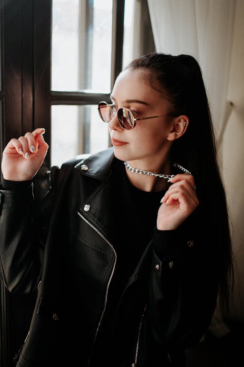 Brunette Woman Wearing Sunglasses, Silver Necklace and Black Leather Jacket Posing by a Window