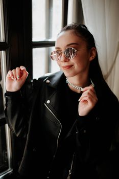 Stylish woman in leather jacket striking a pose by a sunlit window.