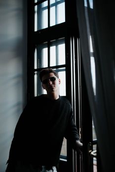 Artistic portrait of a man standing by a window wearing sunglasses, captured in low light for a moody effect.