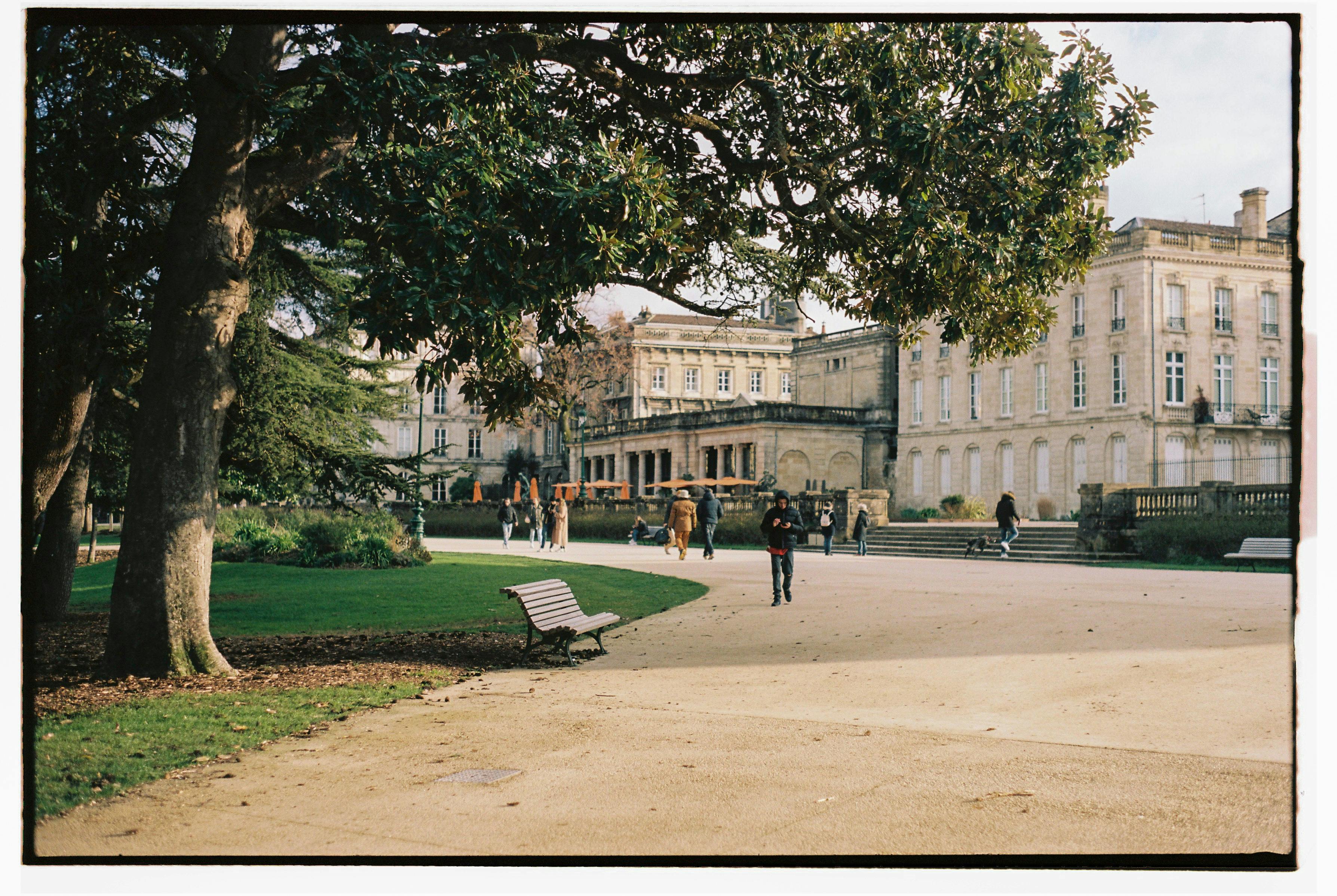 A picturesque park in Bordeaux with people walking, benches, and a historic building.