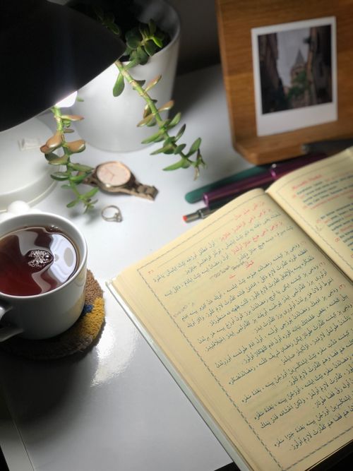 Free A cozy setup with an open Arabic book, tea, lamp, and plant, perfect for studying at night. Stock Photo